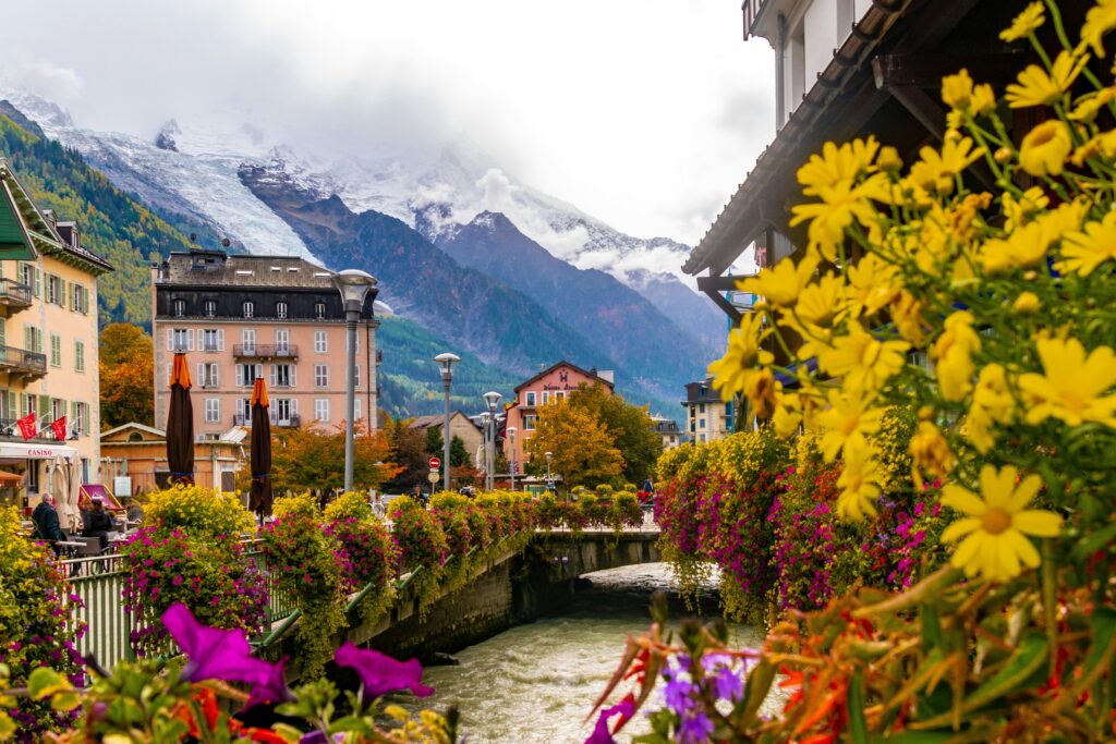 Colorful flowers along a bridge in Chamonix, France, with alpine buildings and the snow-covered Mont Blanc massif rising behind the town.