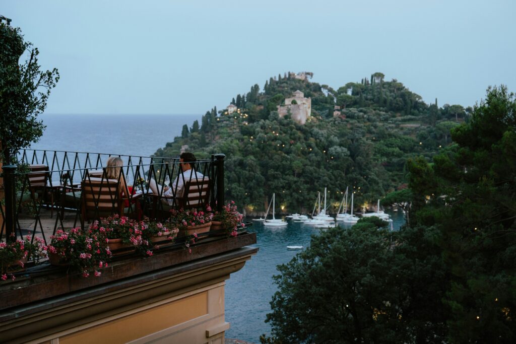Couple seated on a flower-lined terrace overlooking Portofino harbor at dusk, with sailboats and a hillside villa in the background.