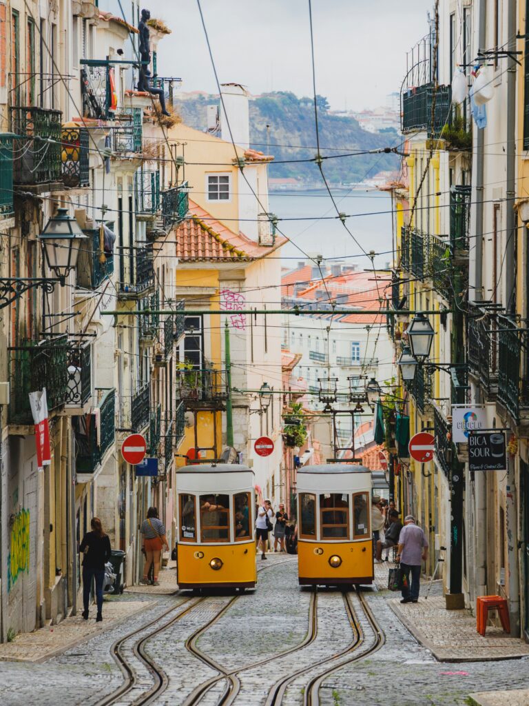 Yellow tram on a narrow cobblestone street in Lisbon's Bairro Alto, with the Tagus River visible in the distance
