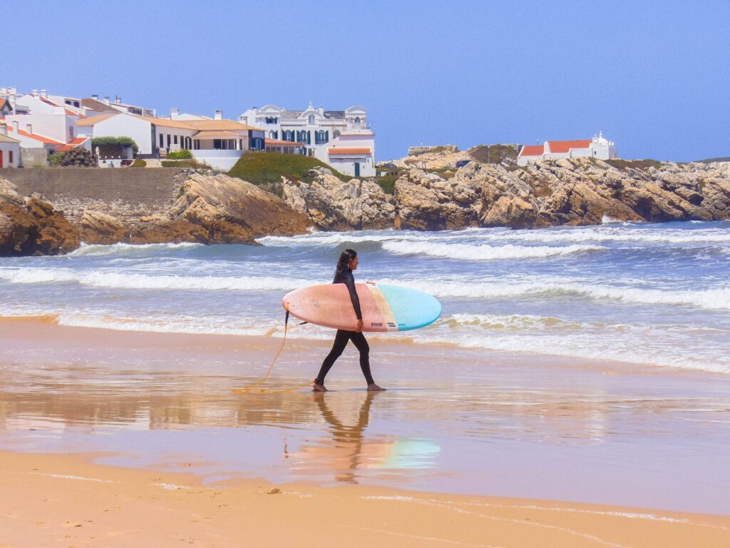 Person carrying a surfboard along a beach in Portugal.